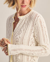 A woman with short blonde hair wears the Daisy Long Sleeve Crew Cardi in Ivory by Velvet by Graham & Spencer, featuring delicate openwork and yellow trim. She accessorizes with rings and a necklace, her face partially visible.