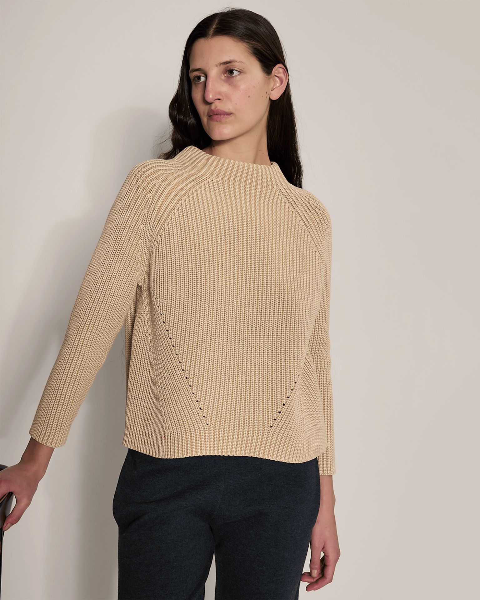 A person with long brown hair wears the Demylee Daphne Washed Cotton Sweater in Sandstone and dark pants, standing against a plain light wall and looking slightly to the side with one hand resting on a surface.