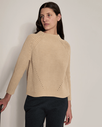 A person with long brown hair wears the Demylee Daphne Washed Cotton Sweater in Sandstone and dark pants, standing against a plain light wall and looking slightly to the side with one hand resting on a surface.