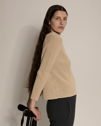 A person with long brown hair stands sideways, wearing the Demylee Daphne Washed Cotton Sweater in Sandstone and dark pants, resting one hand on a black wooden chair against a neutral background.