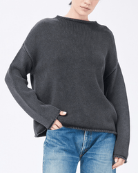 A person stands against a plain light background wearing the Demylee Lamis Washed Cotton Sweater in Black with relaxed hands by their sides. Their face is partially visible and they pair the sweater with faded blue jeans.