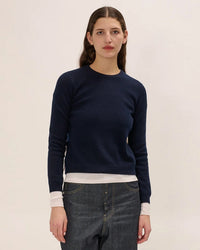 A person with long brown hair, wearing the Demylee Lulu Sweater in Navy over a white shirt and blue jeans, stands against an off-white background, looking straight ahead with a neutral expression.