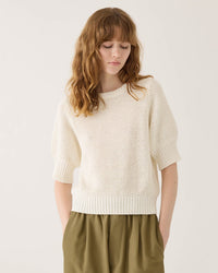 A woman with wavy, light brown hair wears the Demylee Sherry Cotton Top in White and olive green pants, standing against a plain light background and looking down with her hands in her pockets.