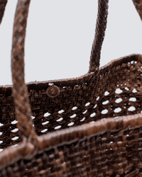 Close-up of Dragon Diffusion's Cannage Max in Dark Brown, a handwoven leather tote with two handles and a small round metal tag by the rim, highlighting its detailed texture against a plain light background.