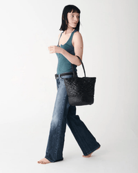A woman with straight black hair, in a teal tank top and blue jeans, walks barefoot carrying the Dragon Diffusion Corso Bucket Bag in Black, glancing to her left against a plain white background.