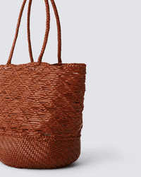 The Corso Bucket Bag in Tan by Dragon Diffusion is a brown woven leather bag with dual handles, showcasing a crisscross texture on top and a tighter weave below, set against a plain light gray background.