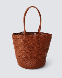 The Corso Bucket Bag in Tan by Dragon Diffusion is a brown woven straw tote with two handles, a textured pattern, and a round base, shown against a plain light gray background.