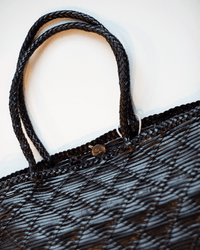 Close-up of the Dragon Diffusion NS Corso Bag in Black, showcasing two braided handles, a textured woven leather surface, and a small round metallic charm near the top edge, set against a white background.
