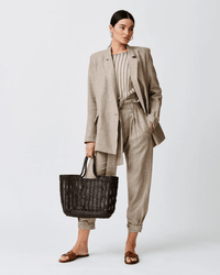 A woman stands against a plain background, wearing a beige suit with pleated trousers, a striped top, brown sandals, and carrying the Dragon Diffusion Window Basket in Dark Brown. Her hair is tied back and she wears hoop earrings.