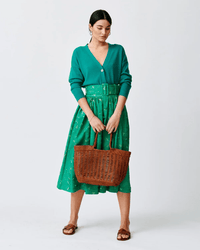 A woman in a green cardigan and skirt stands against a white background, holding the Dragon Diffusion Window Basket in Tan. She wears brown sandals, with her hair tied back.