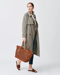 A woman in a long plaid trench coat, blue jeans, and brown slide sandals carries the Dragon Diffusion Window Shopper in Tan with a braided handle. She stands against a plain light background, looking to the side.