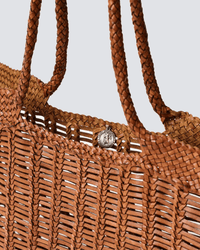 Close-up of the Window Shopper in Tan by Dragon Diffusion, a hand-crafted leather tote with a braided handle and a silver coin-like charm near the opening, set against a plain light gray background.