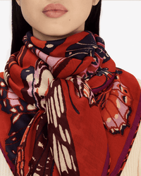 A woman wears the Inoui Editions Square / Carre 130 Papillons in Red scarf wrapped around her neck, shown from the shoulders up against a plain background.