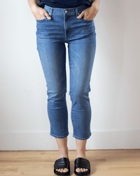 A person stands indoors against a white wall, wearing Edwin's Bree in Dreamland straight leg jeans, a dark top, and black slide sandals with hands in their pockets. Their head and upper torso are not visible.