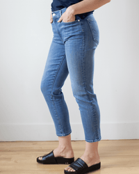 Wearing Edwin's Bree in Dreamland jeans, a dark top, and black open-toe slide sandals, a person stands on a wooden floor with one hand in their pocket against a plain white wall.