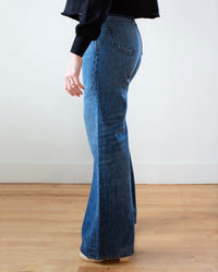 A person stands indoors on a wooden floor, wearing Edwin’s Iris Palazzo in Milton and a black long-sleeve top. The side view highlights the wide-leg fit and flattering shape of the jeans.