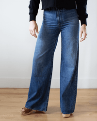 Wearing Edwin's Iris Palazzo in Milton—high-waisted, wide-leg cotton denim jeans—with a black sweater. The lower half is visible, standing on a wooden floor against a white wall.