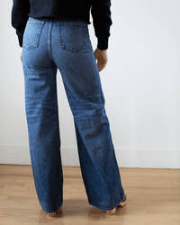 A person in Edwin’s Iris Palazzo in Milton high-waisted wide-leg jeans and a black long-sleeve top stands barefoot on a wooden floor, facing away from the camera against a white wall.