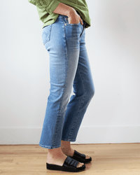 A person indoors wears Edwin Lark Ankle in Paradigm jeans, a green long-sleeve shirt with rolled-up sleeves, and black open-toe slide sandals. They have their hands in their pockets, standing by a white wall and wooden floor.