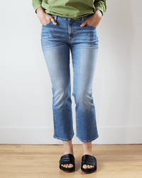 Person wearing Edwin’s Lark Ankle in Paradigm jeans (light blue, subtle flare leg), a green long-sleeve shirt, and black slide sandals, standing indoors on a wooden floor with hands in pockets; head not visible.