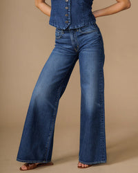 A person showcases the lower half of their body in Edwin’s Marli Super Wide Leg in Rialto jeans, paired with a matching denim top, standing against a beige background with one hand on their hip.