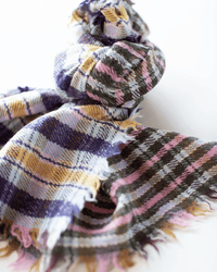 A close-up of the Épice Check Blanket Scarf in Pink, an oversized 100% wool scarf with frayed edges and plaid patterns in purple, pink, mustard yellow, white, and brown, loosely tied against a white background.