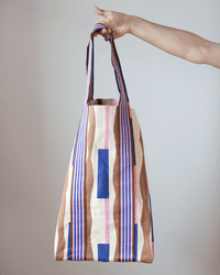 A hand holds the Épice Cotton Bag in Lavender - Large, a summer-ready tote with vertical stripes in brown, pink, blue, and cream, and solid blue accents on a light canvas background.