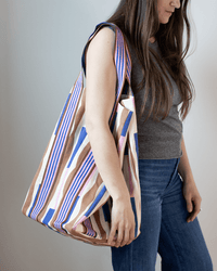 A woman with long brown hair, wearing a gray sleeveless top and blue jeans, carries the Épice Cotton Bag in Lavender - Large over her shoulder. The spacious summer tote features vibrant blue, pink, tan, and white stripes.