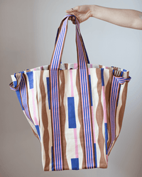 A hand holds the Épice Cotton Bag in Lavender - Large, featuring hand screen printed wavy stripes and wide handles—an oversized cotton canvas tote, ideal for summer, shown against a neutral background.