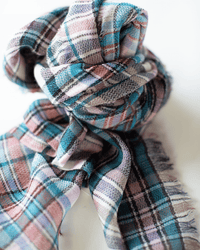 A close-up of the Épice Small Square Check Scarf in Turquoise, featuring a textured hand-woven wool fabric with frayed edges, shown loosely knotted on a white background.