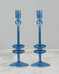 The Sapphire Glass Candlesticks Set of 2 by etúHOME, crafted from recycled glass with round bases and decorative rings, elegantly grace the marble surface, adding a refined touch to home decor.