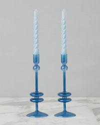 The Sapphire Glass Candlesticks Set of 2 by etúHOME elegantly holds spiral blue taper candles on a sleek marble surface. Crafted from recycled glass, they offer a sustainable touch of beauty to your home decor.