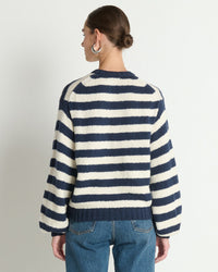 A person with brown hair in a low bun, wearing large hoop earrings, blue jeans, and the Nation LTD Tessa Cardigan in Marine Stripe, stands facing away from the camera against a plain white wall.