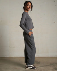 A woman stands sideways against a plain, light wall, wearing the perfectwhitetee Fiona L/S Recycled V Neck Charcoal crop top with matching wide-leg pants and black Nike sneakers featuring white swooshes.