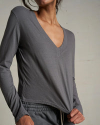 A person stands against a plain light background, wearing the perfectwhitetee Fiona L/S Recycled V Neck Charcoal top and matching drawstring pants. The look features recycled cotton in a loose fit; their head is partially out of frame.