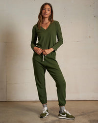 A woman with long hair stands indoors against a plain wall, wearing the perfectwhitetee Fiona Long Sleeve Recycled V Neck in Fern Green and green sneakers with white soles, her hands resting in her pockets.