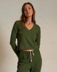 A woman wears the perfectwhitetee Fiona Long Sleeve Recycled V Neck in Fern Green, paired with drawstring pants, standing against a plain light wall and looking at the camera with a relaxed expression.
