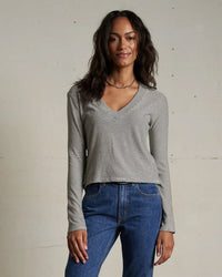 A woman with wavy dark hair smiles softly, wearing the perfectwhitetee Fiona Stripe Jersey V Neck L/S in Ivory/Black and blue jeans, standing against a plain beige wall.