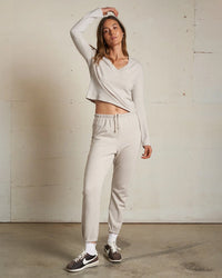 A woman poses indoors against a light wall, wearing perfectwhitetee’s Johnny French Terry Easy Sweatpant in Silver, a matching beige crop top, white socks, and black sneakers. She raises one arm and looks up in stylish loungewear.