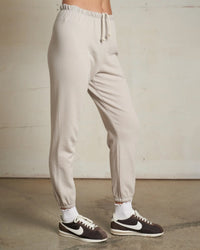 Wearing the perfectwhitetee Johnny French Terry Easy Sweatpant in Silver with white socks and black-and-white Nike sneakers, standing on a concrete floor against an off-white wall. Only the lower half is visible—modern loungewear style.