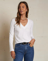 A woman with long brown hair wears the perfectwhitetee Fiona Long Sleeve Recycled V Neck in White, paired with blue jeans and a black belt. She stands before a neutral wall, gazing to the side with a relaxed expression.