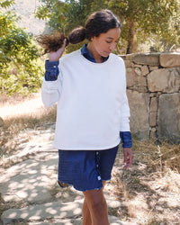 A woman walks outdoors on a stone path, wearing the Frank & Eileen Anna Long Sleeve Fleece Capelet in White over a blue denim shirt and shorts. She holds her curly hair, with trees and a stone wall behind her.