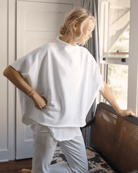 A woman stands by a window, smiling in soft natural light as she wears the Frank & Eileen Audrey Funnel Neck Capelet in White, paired with white pants and resting a hand on a wooden chair with a woven back.