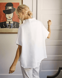A woman with blonde hair in a loose bun stands with her back to the camera, wearing Frank & Eileen's Audrey Funnel Neck Capelet in White and white pants in a bright room, beside a colorful framed painting on the wall.