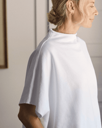 A woman with blonde hair in a low bun wears the Frank & Eileen Audrey Funnel Neck Capelet in White, standing sideways in a softly lit room. Only her upper body and part of her face are visible.