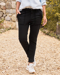 A person stands on a gravel path wearing Frank & Eileen’s Blackrock Utility Sweatpant in Black, styled with a light blue rolled-sleeve button-up and white sneakers. Their hands are in their pockets; only their body from the shoulders down is shown.