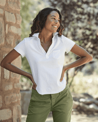 A woman stands outdoors by a stone wall, smiling with her hands on her hips. She wears green pants and the Frank & Eileen Charlotte S/S Polo in White. Blurred greenery can be seen in the background.
