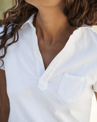A person with wavy brown hair wears the Frank & Eileen Charlotte S/S Polo in White, showing only their lower face, neck, and upper torso against a softly blurred background.