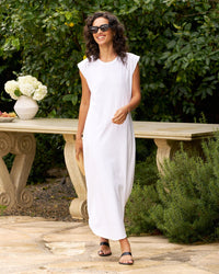 A woman in the Frank & Eileen Cory Vintage Muscle Tee Dress in White smiles outdoors on a stone patio, wearing black sandals and sunglasses near a table with white flowers, surrounded by greenery.