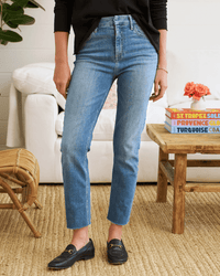 A person stands indoors wearing Frank & Eileen’s Derry Pant in 2001 Classic Blue Wash, a black long-sleeve top, and black loafers. Nearby are a white armchair, wicker stool, and wooden table with books and flowers.